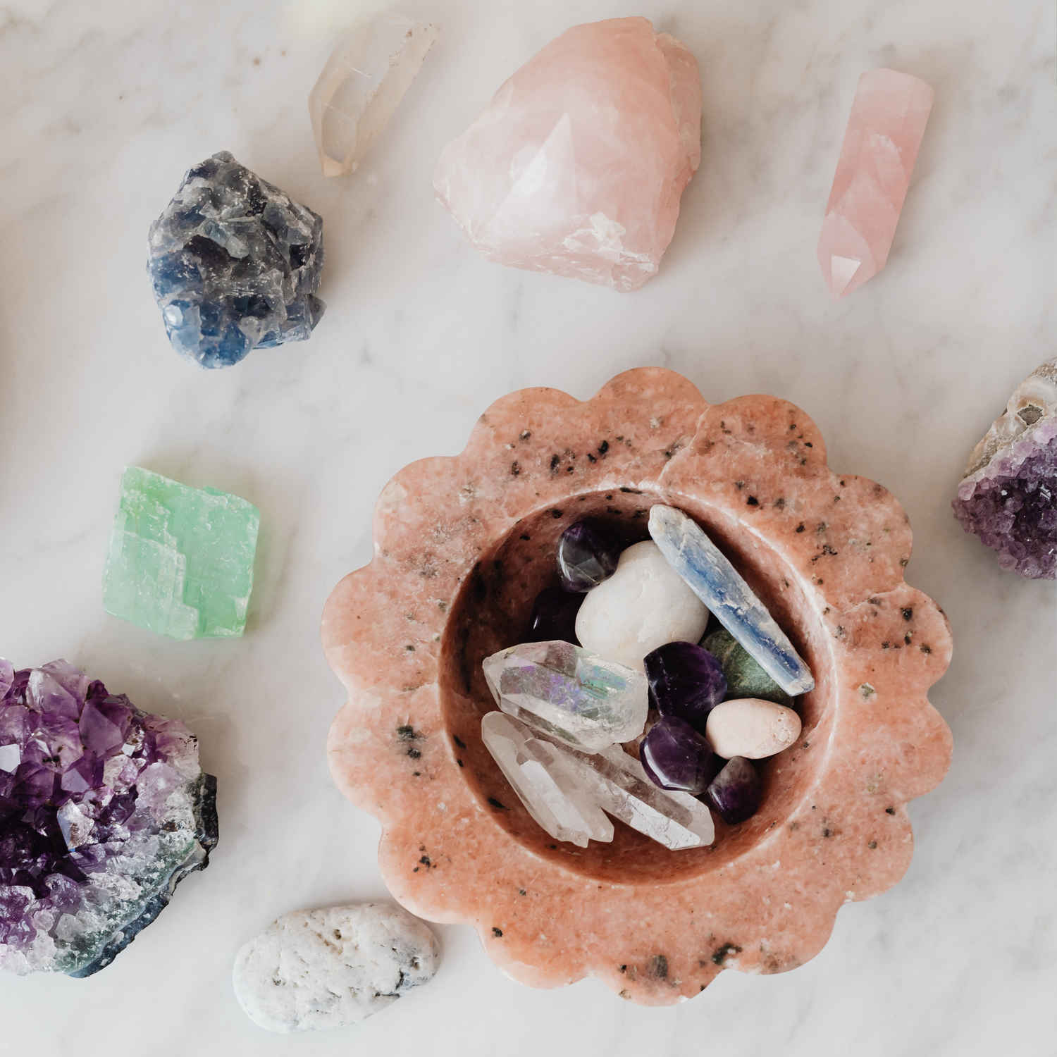 A flat lay of healing crystals arranged on a white marble surface. The centerpiece is a pink flower-shaped stone bowl containing various polished crystals, including clear quartz, amethyst, and kyanite. Surrounding the bowl are unpolished stones like rose quartz, green fluorite, amethyst clusters, and raw blue celestite, representing energy balancing and holistic wellness.