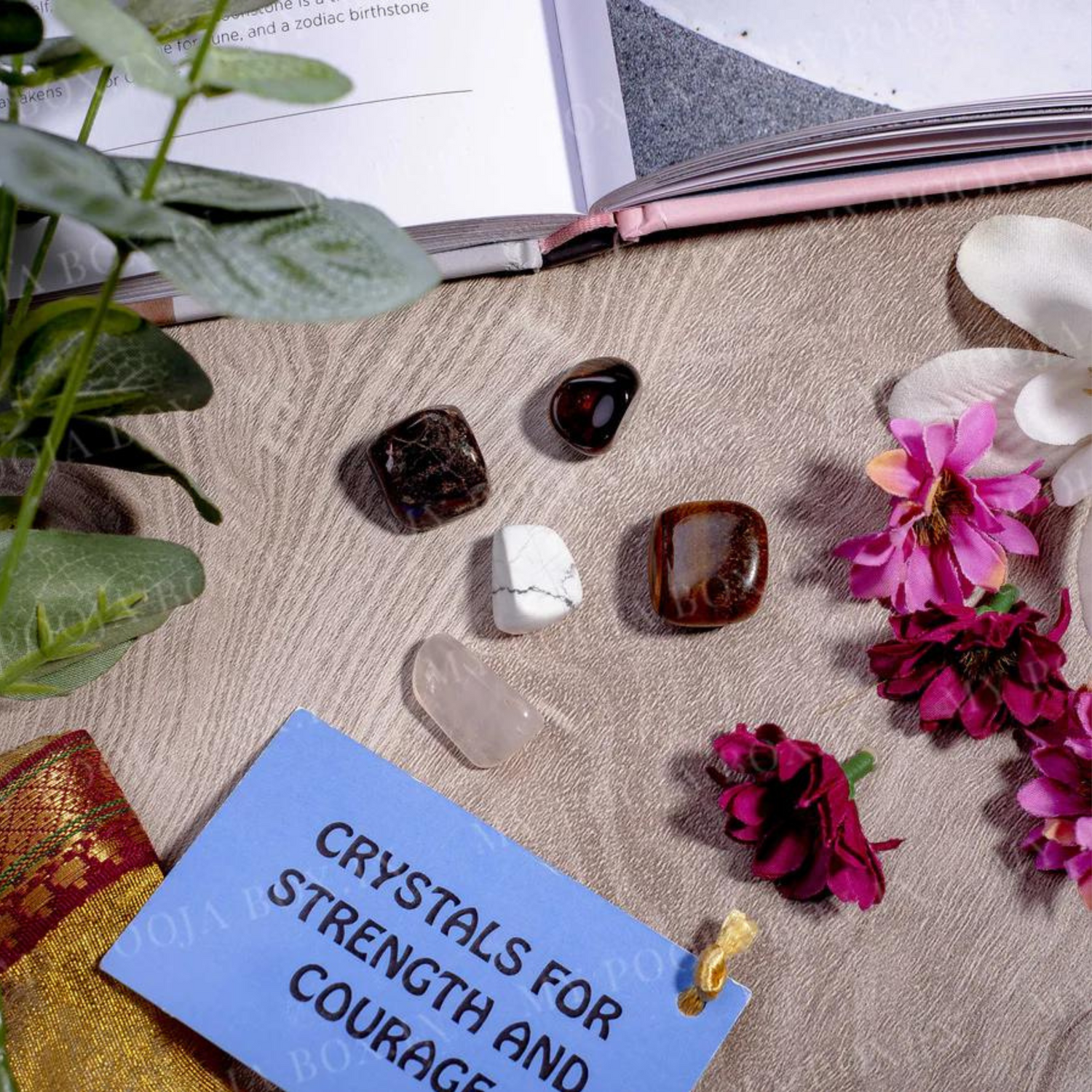 A flat lay of crystals for strength and courage displayed on a textured beige fabric. Featured stones include tiger's eye, garnet, howlite, and rose quartz. Surrounding the crystals are vibrant pink and white flowers, a sprig of greenery, an open book, and a blue tag labeled 'CRYSTALS FOR STRENGTH AND COURAGE.' A touch of gold fabric adds a decorative accent.