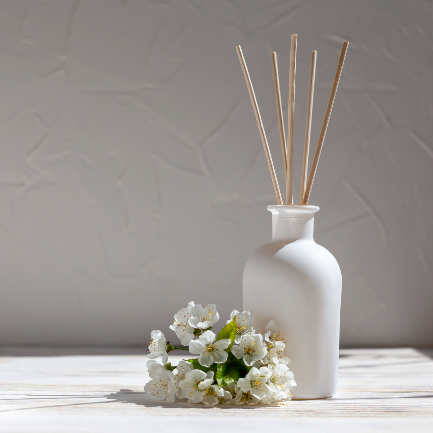 A minimalist arrangement featuring a white ceramic diffuser bottle with wooden reed sticks placed on a light wooden surface. Beside the diffuser is a small cluster of delicate white blossoms with green leaves. The backgrou